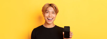 Close-up of stylish handsome korean guy showing smartphone screen and smiling pleased, recommend mobile app, standing over yellow backgroundの写真素材