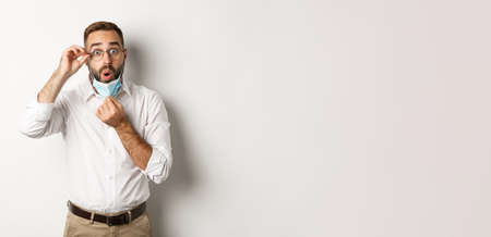 Covid-19, social distancing and quarantine concept. Impressed business man take off medical mask, looking surprised, standing over white backgroundの写真素材