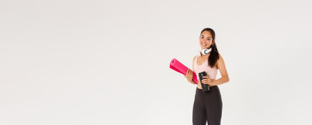 Full length of smiling slim and beautiful asian female athlete, girl carry fitness exercise rubber mat and water bottle, going for yoga classes or workout in gym with own equipment, white backgroundの写真素材