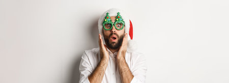 Adult man celebrating winter holidays, wearing christmas party glasses and santa hat, looking surprised at camera, standing over white backgroundの写真素材