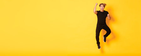 Full-length of happy attractive asian guy in black clothes jumping and celebrating victory, achieve...