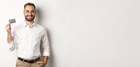 Handsome businessman showing his credit card, looking satisfied, standing over white backgroundの写真素材