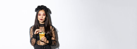 Confident smiling asian woman looking satisfied, holding sweets and wearing witch costume on...