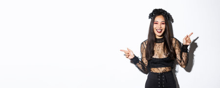 Beautiful happy young woman celebrating halloween in black lace dress, gothic style makeup, pointing fingers sideways, showing two banners or products, standing over white backgroundの写真素材