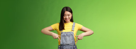Studio shot easygoing cute asian female introduce new promo, pointing looking down amused pleasantly smiling, gladly show interesting thing, stand green background happy excitedの写真素材
