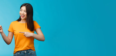 Friendly enthusiastic asian girl curiously looking sideways, pointing left interesting object, grinning joyfully discuss new opened store, stand blue background carefree lively talkingの写真素材