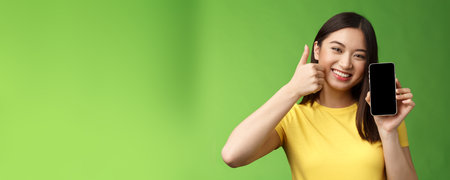 Close-up satisfied cute asian girl give positive feedback, thumb-up showing smartphone screen, introduce awesome new app, recommend good application, bragging beating score in gameの写真素材