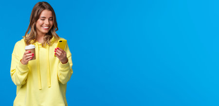 Carefree joyful european female in yellow hoodie, holding take-away coffee and browsing internet in...