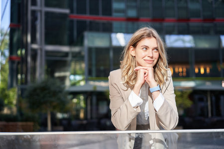 Beautiful young woman in beige suit, standing near office buildings in city center, smiling and looking dreamyの写真素材