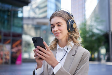 Portrait of smiling young businesswoman listening music in headphones and using mobile phone while on a busy city streetの写真素材