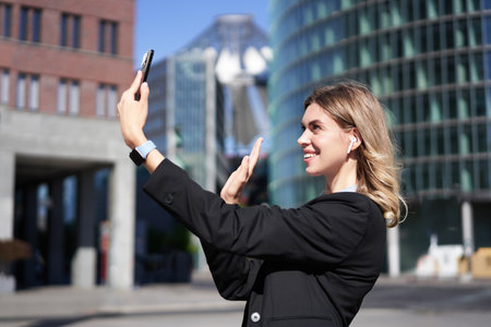 Portrait of businesswoman wave her hand at mobile phone camera, waves hand during video chat, stands in suit in city center outdoorsの写真素材