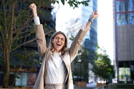 Happy businesswoman scream of joy, raise hands up in celebration, cheering gesture, achieve goal and triumphing on city streetの写真素材