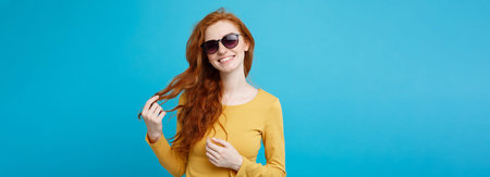 Travel concept - Close up Portrait young beautiful attractive ginger red hair girl with trendy sunglass smiling. Blue Pastel Background. Copy space.の写真素材