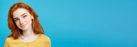Lifestyle concept - Portrait of cheerful happy ginger red hair girl enjoy listening to music with headphones joyful smiling to camera. Isolated on Blue Pastel Background. Copy space.の写真素材