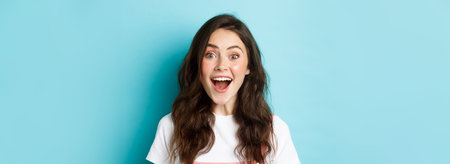 Close up portrait of surprised and happy young woman checking out awesome news, looking amazed with opened mouth, fascinated face, standing over blue backgroundの写真素材