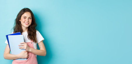 Cute smiling girl student holding notebooks and looking cheerful at camera, studying in college or university, standing over blue backgroundの写真素材