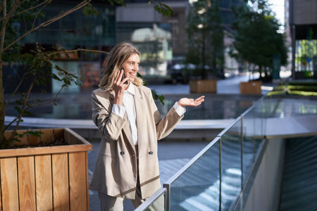 Beautiful young businesswoman in suit talks on mobile phone on street. Corporate woman calls someone on phone while stands outsideの写真素材