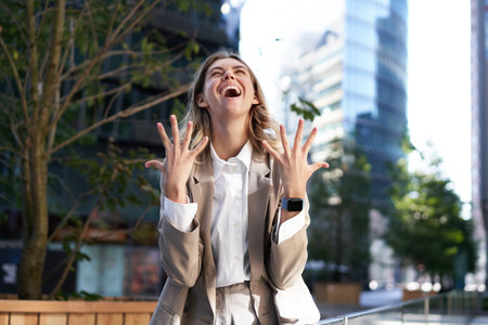 Enthusiastic businesswoman rejoicing, celebrating and feeling happy, express pure joy, standing on street in beige suitの写真素材