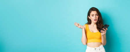 Technology and lifestyle concept. Glamour girl looks confused after reading smartphone screen, staring puzzled at camera, standing against blue backgroundの写真素材