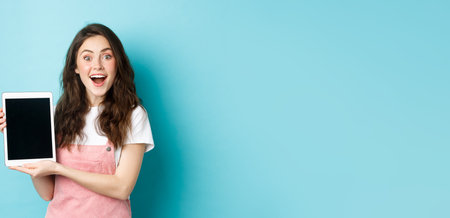 Amused pretty girl demonstrate blank digital tablet screen and smiling excited at camera, showing awesome promo offer, standing against blue backgroundの写真素材