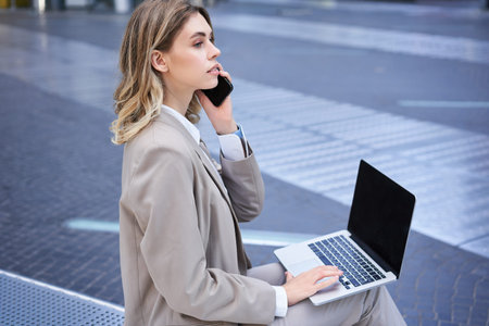 Young corporate woman sitting in city centre with laptop and talking on mobile phoneの写真素材