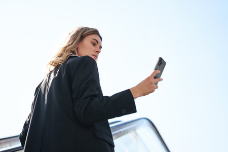 Successful businesswoman standing on escalator, looking at mobile phone, reading message on smartphone, view from behindの写真素材