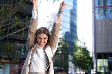 Saleswoman expresses joy and happiness. Businesswoman triumphing on street, raising hands up, celebrating victory, smiling pleasedの写真素材