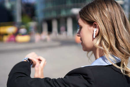 Portrait of busy corporate woman in wireless headphones, looking at time on her digital watch,...