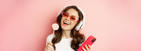 Beautiful female model listening music in headphones, holding lolipop and mobile phone, posing in sunglasses, standing over pink backgroundの写真素材