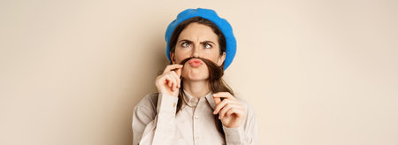Funny beautiful girl fooling around, making moustache out of hair under lips, squinting and...