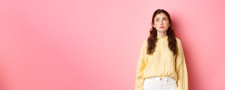 Image of upset and gloomy young woman pulling sad face, looking up with regret or jealousy, standing...