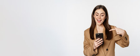 Enthusiastic saleswoman, business woman pointing finger at mobile phone and smiling, showing on cellphone, standing against white backgroundの写真素材