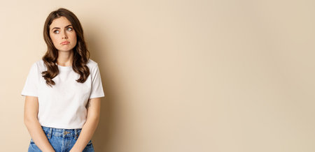 Sad and tired brunette woman looking drained and unamused, standing with pokerface against beige backgroundの写真素材