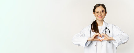 Portrait of smiling doctor, female physician showing heart sign and looking with care, love her patients, standing over white backgroundの写真素材