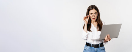 Shocked woman looking at laptop screen confused, stunned about smth on computer, standing over white...