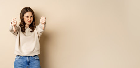 Very bad. Angry woman showing dislike, thumbs down gesture and grimacing from aversion, complaining, negative opinion feedback, beige backgroundの写真素材