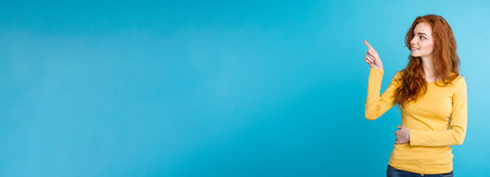 Close up Portrait young beautiful attractive redhair girl happy with something and pointing finger. Blue Pastel Background. Copy spaceの写真素材