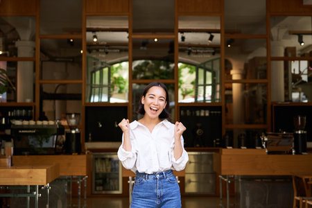 Happy enthusiastic asian girl, business owner triumphing, opening her own restaurant, standing in front of empty cafeの写真素材