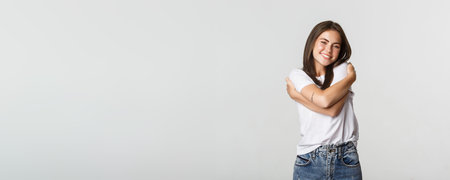 Attractive smiling tender woman hugging herself on white background.