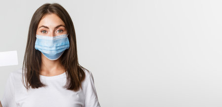 Covid-19, health and social distancing concept. Close-up of pretty brunette girl in medical mask, showing credit cardの写真素材