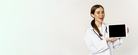 Online medical help, remote appointment. Smiling beautiful woman doctor showing digital tablet screen, demonstrating website logo, standing over white backgroundの写真素材