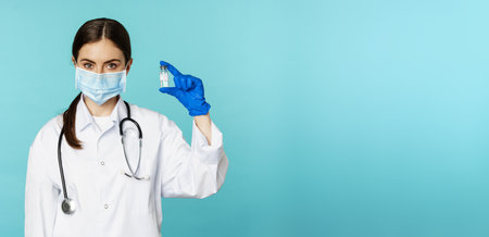 Vaccination campaign. Smiling woman doctor in face medical mask and gloves, showing vaccine from covid, omicron delta variant, standing over torquoise backgroundの写真素材