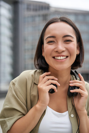 Vertical shot of beautiful asian woman posing with headphones around neck, smiling and laughing,...