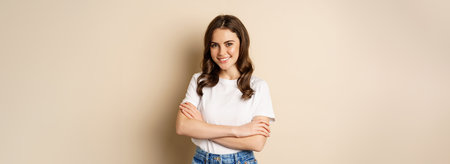 Portrait of beautiful young woman in white t-shirt, smiling and looking happy, posing against beige...