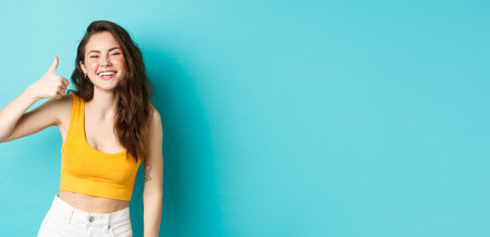 Summer holidays and emotions concept. Cheerful beauty girl with curly hairstyle and cropped top, showing yes thumb up gesture and laughing, standing over blue backgroundの写真素材