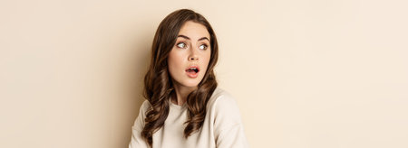 Close up portrait of brunette feminine woman turn back, look behind with surprised face expression, standing against beige backgroundの写真素材