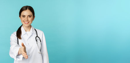Smiling professional woman doctor, therapist extending hand, handshake gesture, greeting patient in medical clinic or hospital, standing over blue backgroundの写真素材