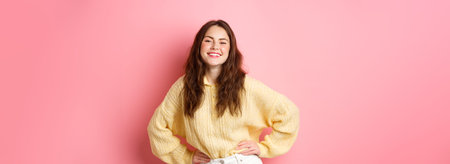 Portrait of beautiful young woman posing and laughing carefree, holding hands on waist, feeling joy...