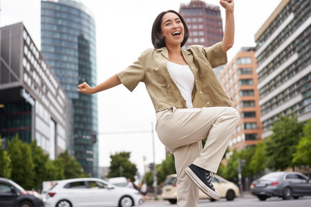 Portrait of dancing asian woman, raising hands up and feeling happy, posing in city, triumphing, feeling joyの写真素材