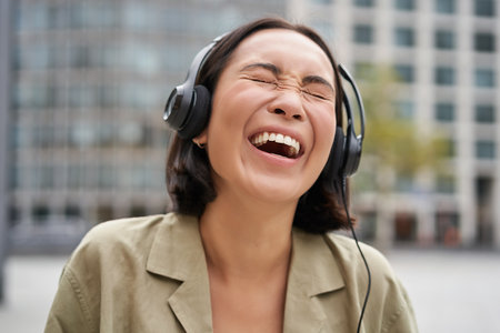 Portrait of happy asian woman in headphones, enjoys music while walking in city, smiling and laughingの写真素材
